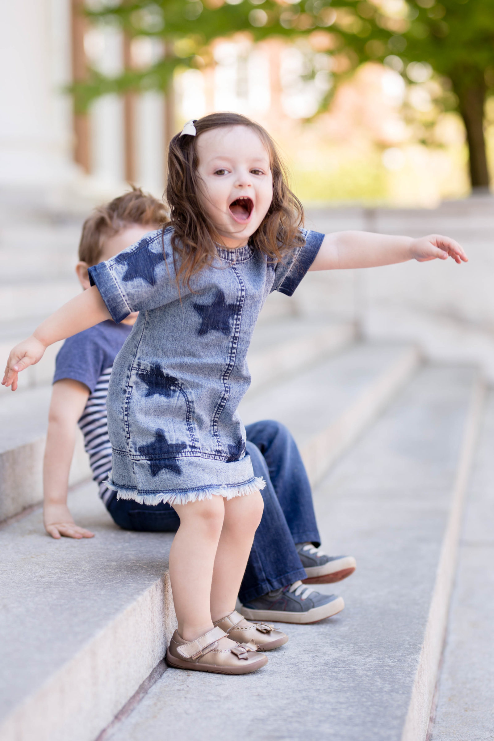 Toddler girl in a denim star dress cheering on stone steps next to her big brother