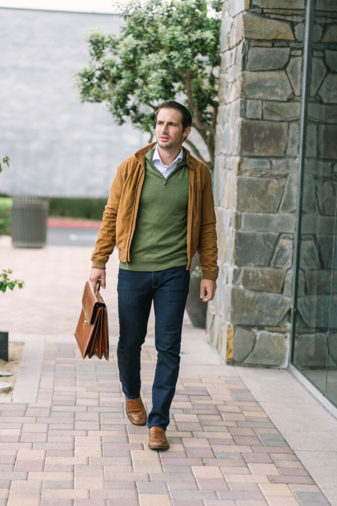 Man in a tan jacket, green sweater and dark jeans for work carrying a briefcase