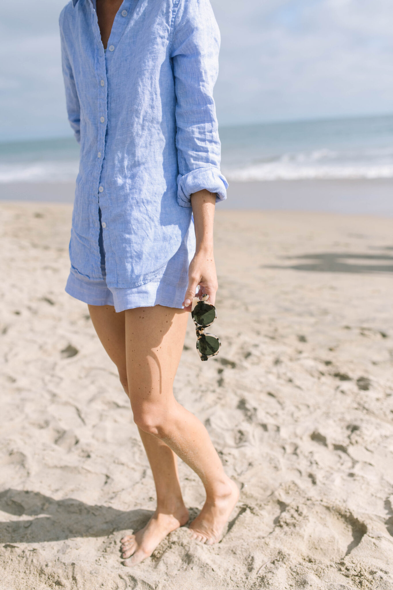 Holding a pair of quince sunglasses on the beach in 2026.